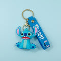A hand holding a blue Stitch character keychain with a matching blue wrist strap and gold metal key ring, against a blurred green hedge background.