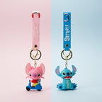 Two Stitch-themed keychains on a light wood surface. One has a pink strap and a pink character charm, and the other has a blue strap and a blue character charm, both with gold keyrings.